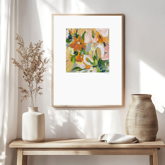 Modern abstract flower wall art print in soft shades of orange, green, warm yellow. Layered flower petals in an expressive style in a square format. Big white border surrounds the design. Framed in a natural frame and hanging on a contemporary neutral living room wall.