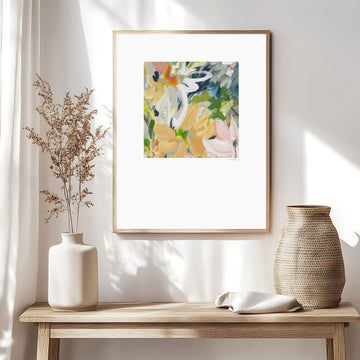Modern abstract flower wall art print in soft shades of orange, green, warm yellow. Layered flower petals in an expressive style in a square format. Big white border surrounds the design. Framed in a natural frame and hanging on a contemporary neutral living room wall.