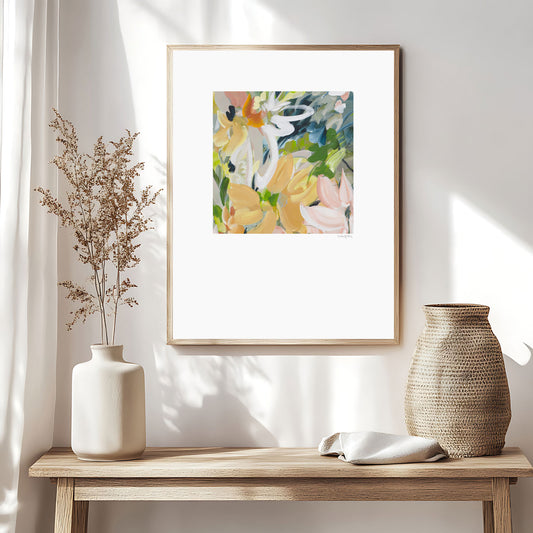 Modern abstract flower wall art print in soft shades of orange, green, warm yellow. Layered flower petals in an expressive style in a square format. Big white border surrounds the design. Framed in a natural frame and hanging on a contemporary neutral living room wall.