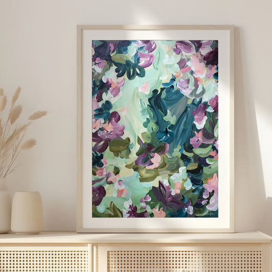 Modern abstract fine art paper print of floating flower petals in spearmint, purple, greens and dark blue. In a natural frame leaning against a wall on a cupboard.