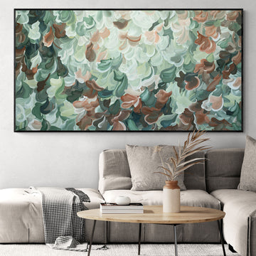 Large green and brown abstract nature painting with a black frame. Hanging in a natural coloured living room with a beige lounge chair.