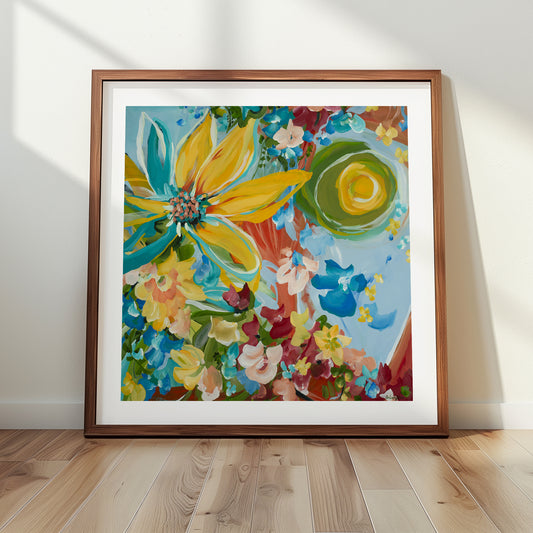 Modern abstract sunflower art print with a big yellow sunflower and smaller red, blue, yellow flowers surrounding it. Colourful wall art print framed in a natural frame and leaning against a wall.
