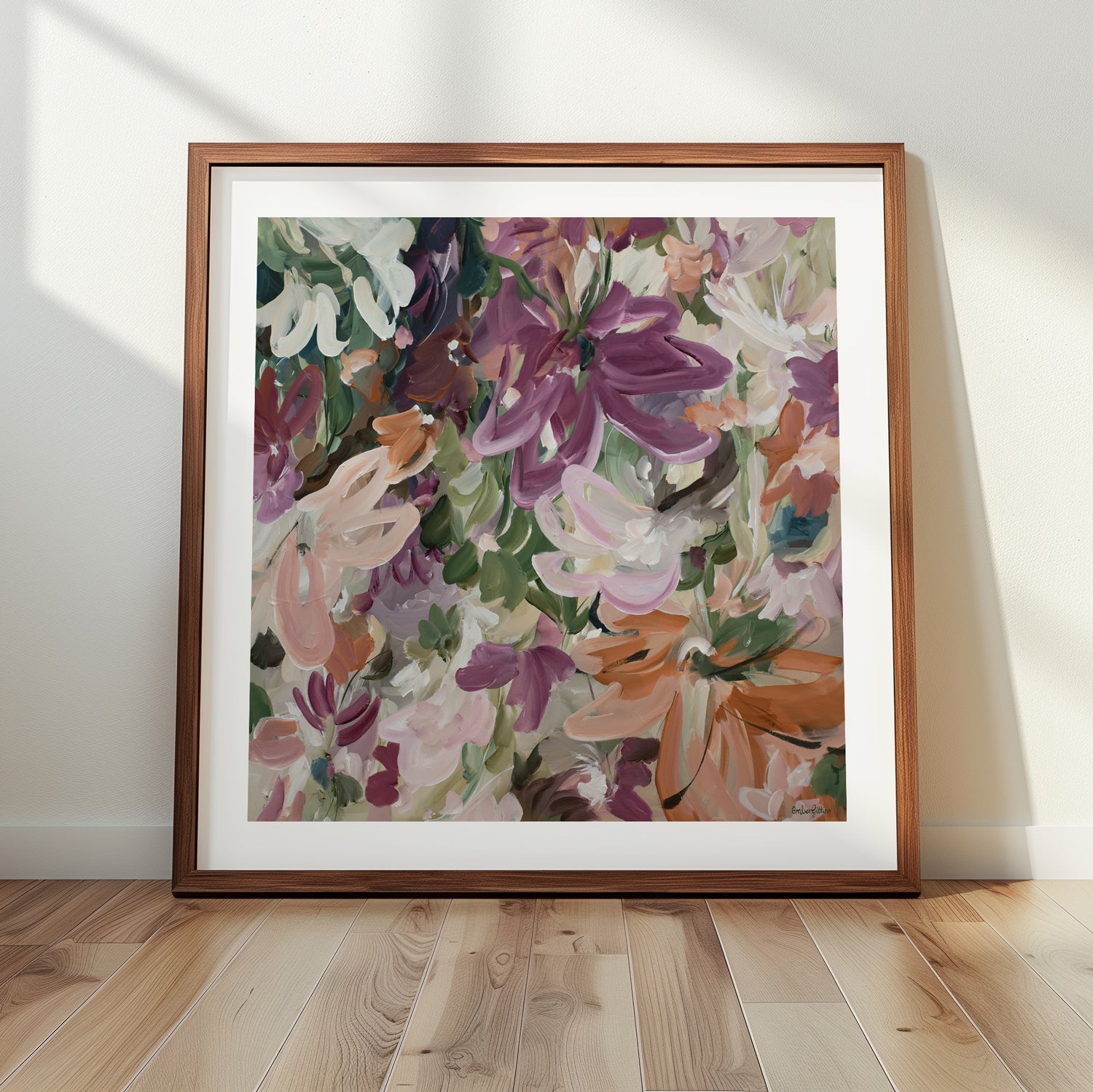 Colourful modern abstract flower fine art wall print in warm tones of red, burgundy, pink, orange and green. Layers of flowing brushstrokes showing linear flower and petal shapes. Framed in a natural frame, leaning against a living room wall.