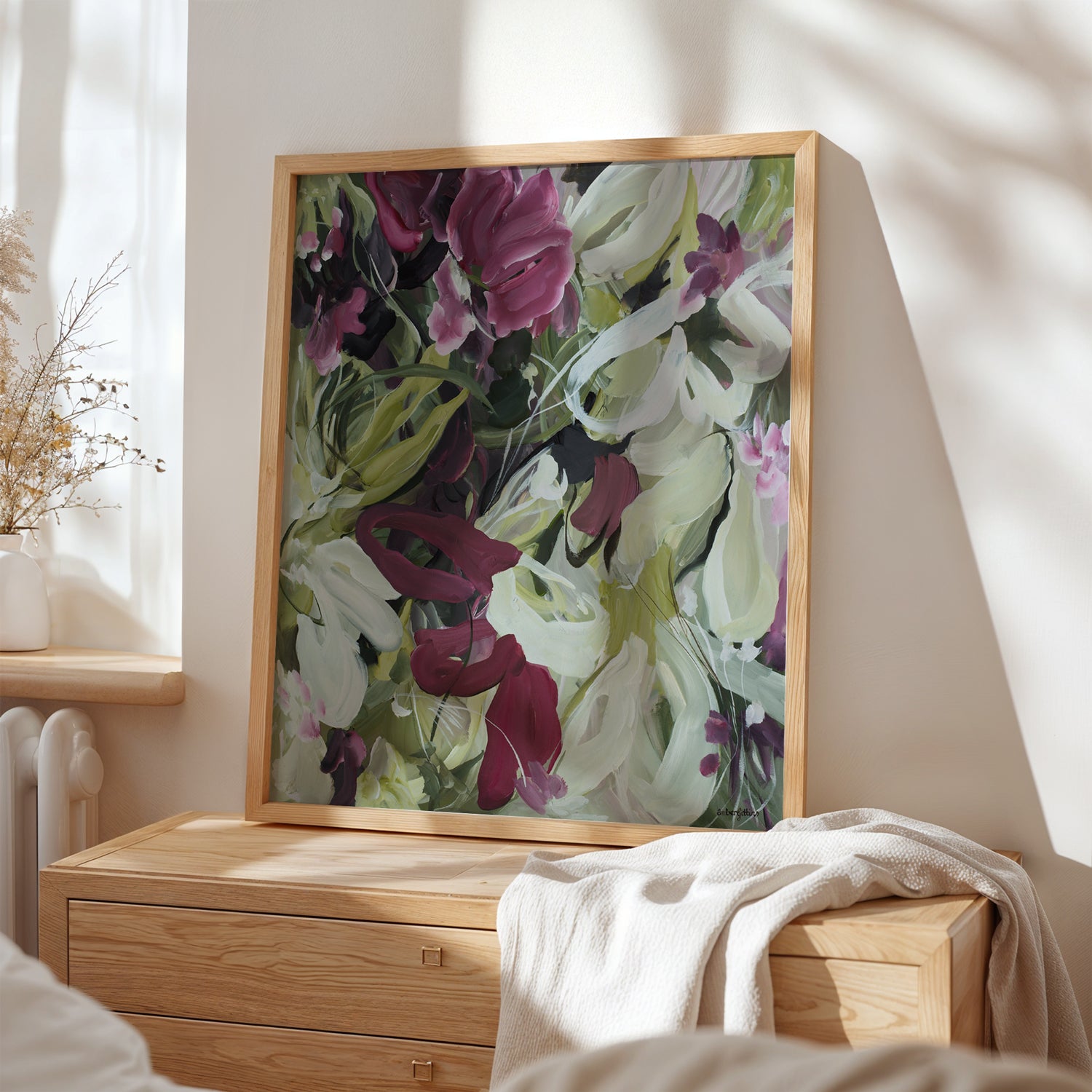 Modern wildflower fine art print in an expressive style in modern colour palette of burgundy, white and green. Abstract floral flowing shapes layered over each other. Framed in a natural frame and leaning against a bedroom wall on a cupboard.
