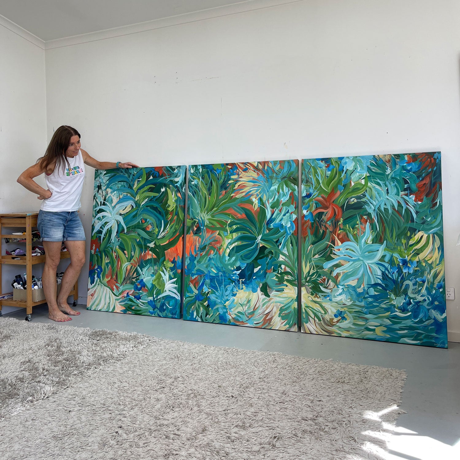 Artist Amber Gittins standing next to three large triptych abstract tropical paintings on a wall