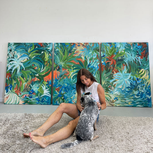 Artist Amber Gittins sitting on the floor of her art studio with a dog in front of a large abstract tropical triptych painting
